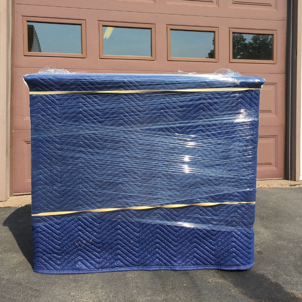 A large piece of furniture fully wrapped in a deep-blue quilted moving blanket and secured with clear stretch wrap, shown in full in bright daylight