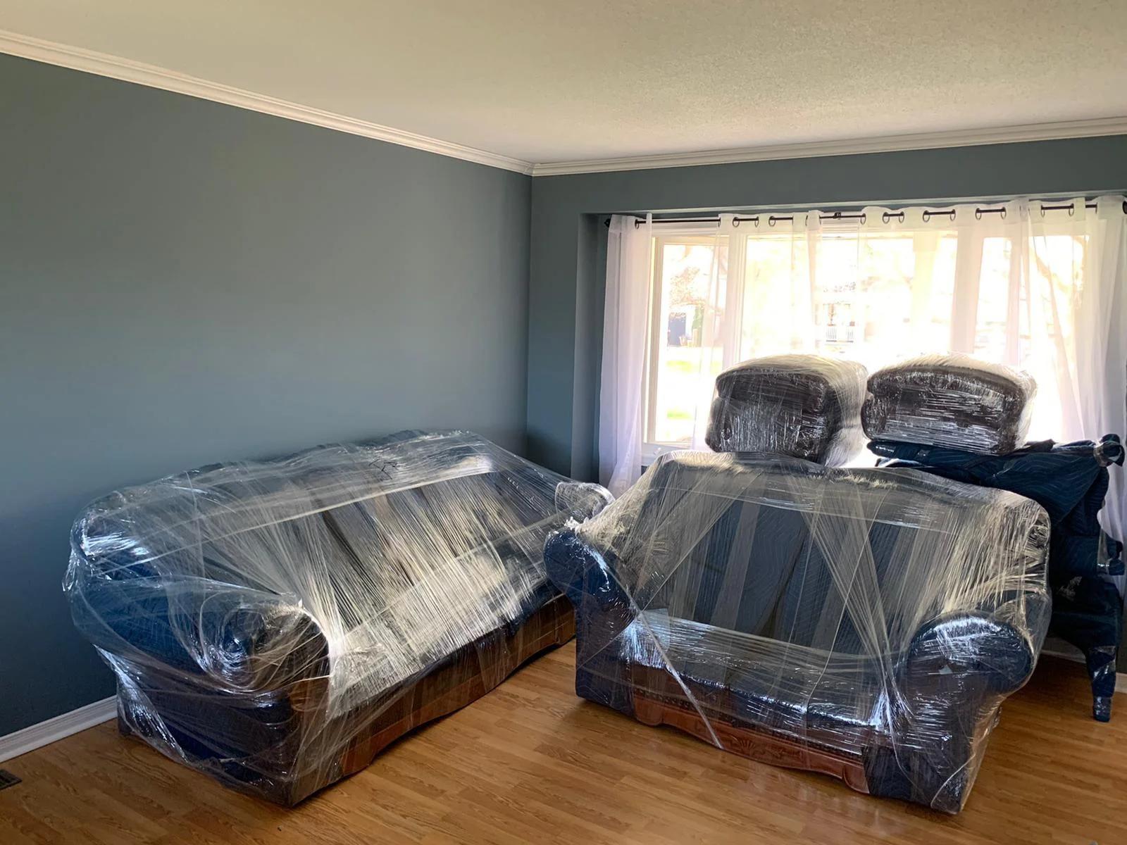 Two sofas fully wrapped in padded moving blankets and sealed with clear stretch wrap, standing in a bright living room with wrapped chairs by the window, shown in full frame and ready to load