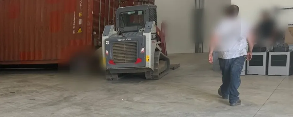 The Flat Rate Movers warehouse and transload operation — crew positioning a red shipping container inside the warehouse with a Toyota forklift and a Takeuchi track loader, with a taped floor zone in the foreground