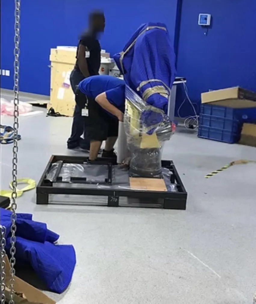 A large industrial robotic arm custom-wrapped in moving blankets and stretch wrap by The Flat Rate Movers, being skid-mounted by crew for transport and storage